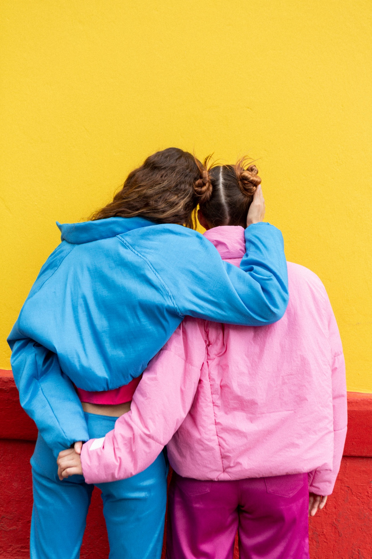 Two people in bright blue and pink jackets with arms around each other against a bold yellow wall, representing the inclusive, joyful Club 'Cure community