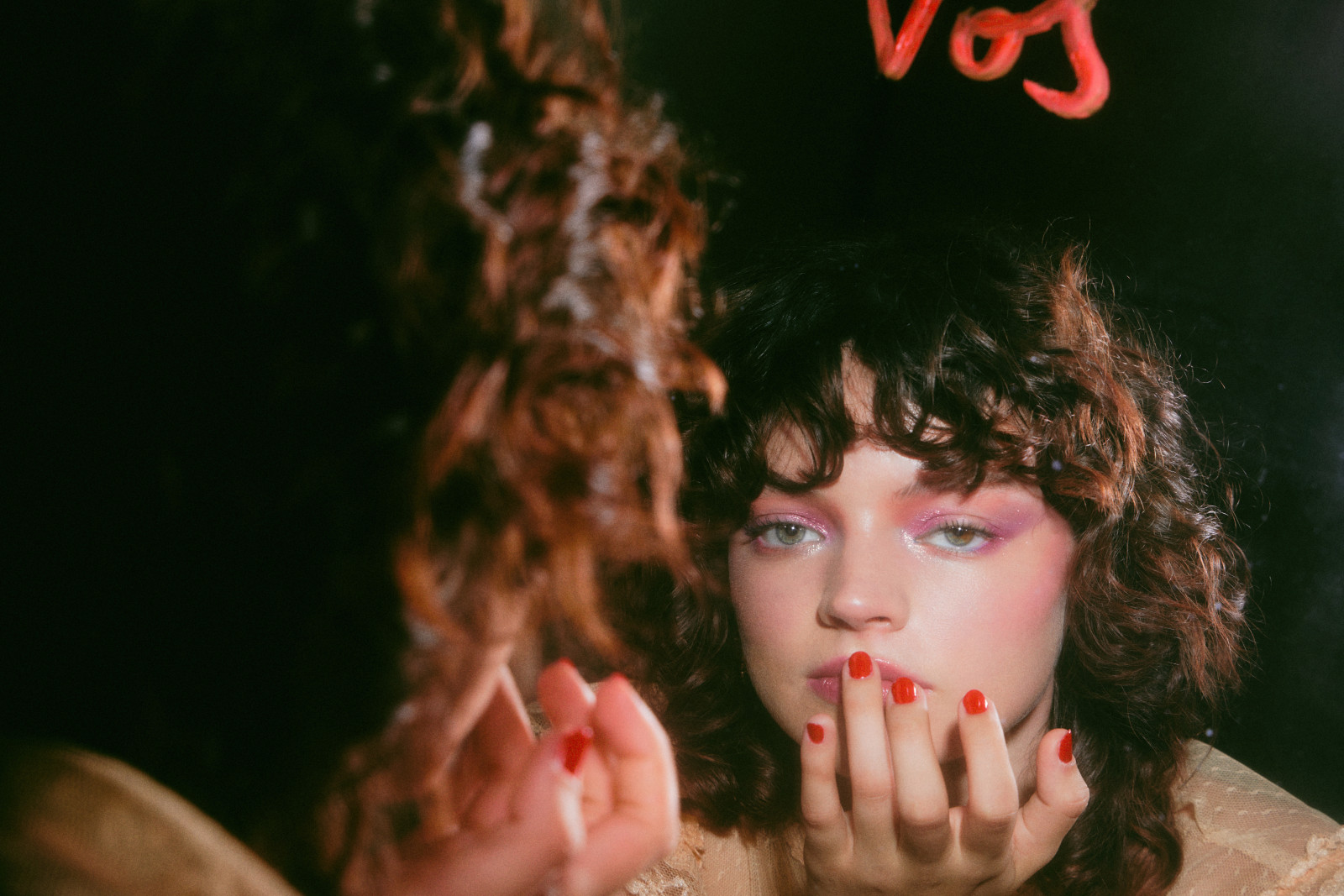 Retro portrait of a model with glossy red nails and pink shadow, reflected in a mirror under warm neon light